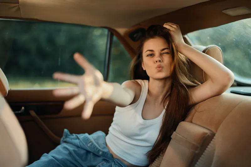 Young woman in casual attire with long hair, playfully posing in a carâ€™s back seat, expressing joy and spontaneity in a vibrant, natural setting.