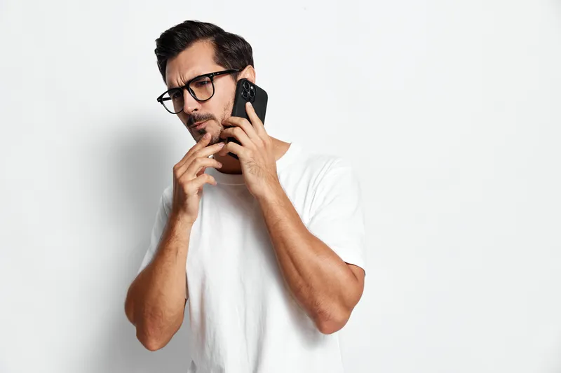 Thoughtful man in white t-shirt wearing glasses, holding smartphone to ear with hand on chin, contemplating while looking away, isolated on plain solid white background.