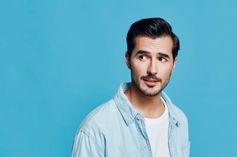 Stylish man looking away with a thoughtful expression, wearing a light blue shirt against a solid vibrant blue background. Concept of contemplation and fashion.