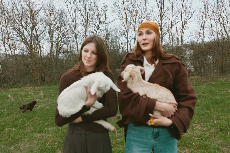 Two women hold a goat kid in an open field, capturing a casual outdoor lifestyle moment with natural light, affectionate expressions, playful animals, and rural charm, everyday bonding.