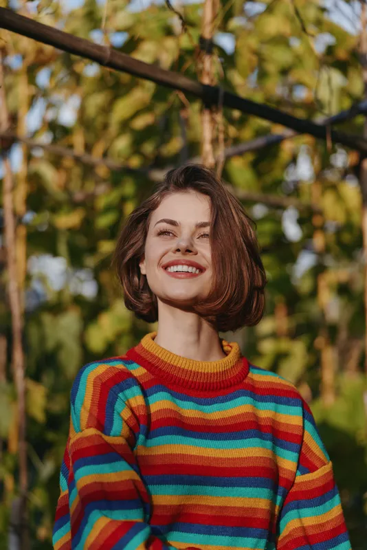 Woman smiling in a colorful striped sweater, short bob hair, outdoor portrait in an autumn garden. Young adult female, casual outfit, joyful expression, lifestyle and fashion image.