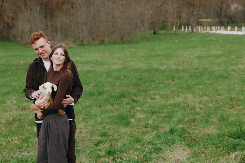 Smiling couple outdoors with a goat kid in a grassy field, embracing warmly. A lifestyle moment displaying family bonding, rural living, and animal companionship in natural light.