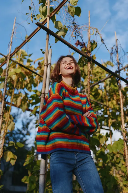 Woman in rainbow sweater with casual jeans and a laugh in a garden by a wooden trellis, folded arms and sunlight, candid portrait of youth, colorful knitwear and relaxed outdoor lifestyle.