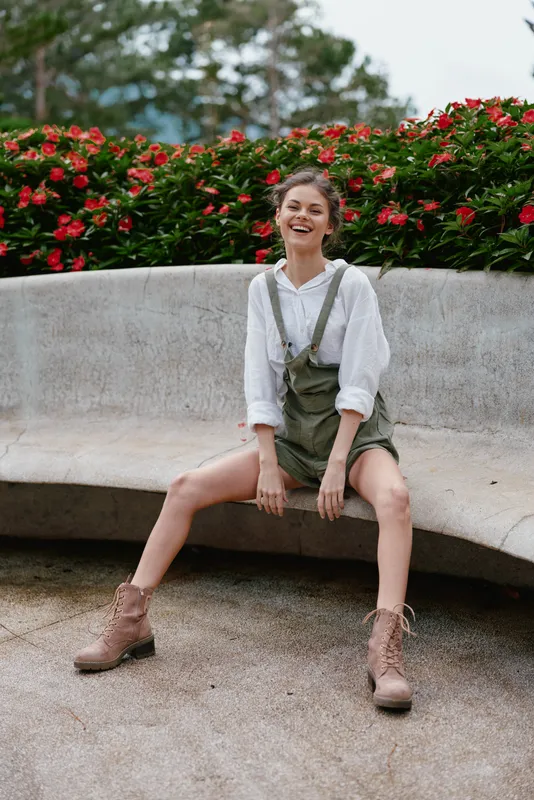 Joyful young woman in overalls sitting on a bench surrounded by vibrant flowers, exuding happiness and confidence in a natural setting