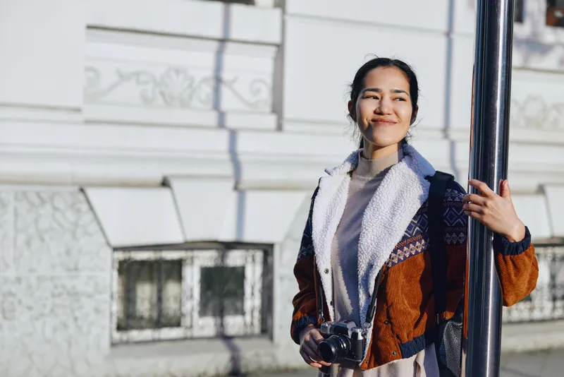 Woman with camera standing near a historic building, wearing a warm coat and scarf, enjoying an urban outdoor moment, ready for street photography and creative storytelling