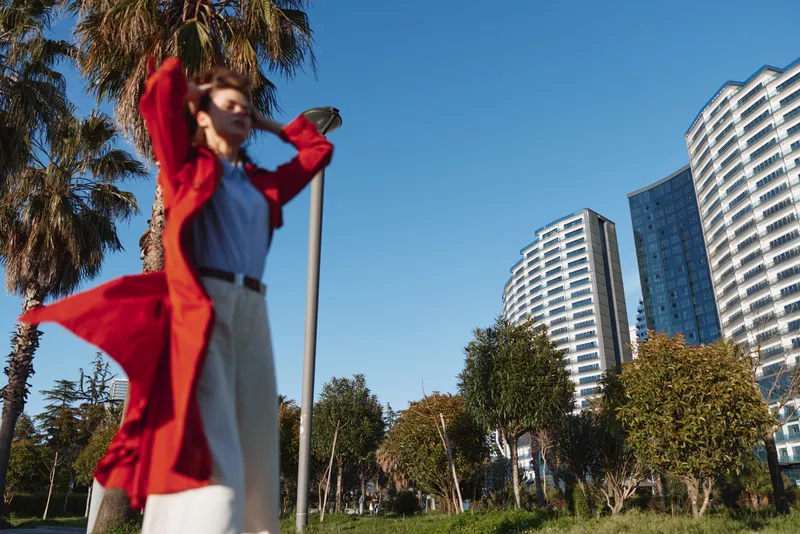 A woman in a red coat stands outdoors near palm trees and modern high rise buildings under a clear blue sky during daytime in an urban park setting.