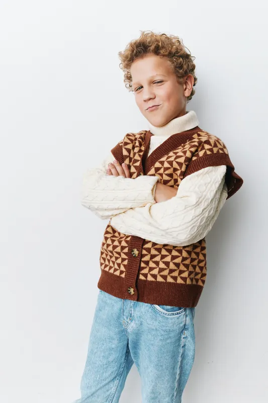 Portrait of a young boy with curly hair wearing a brown geometric knit vest over a cream sweater, arms crossed, a confident smile, and a clean light background.