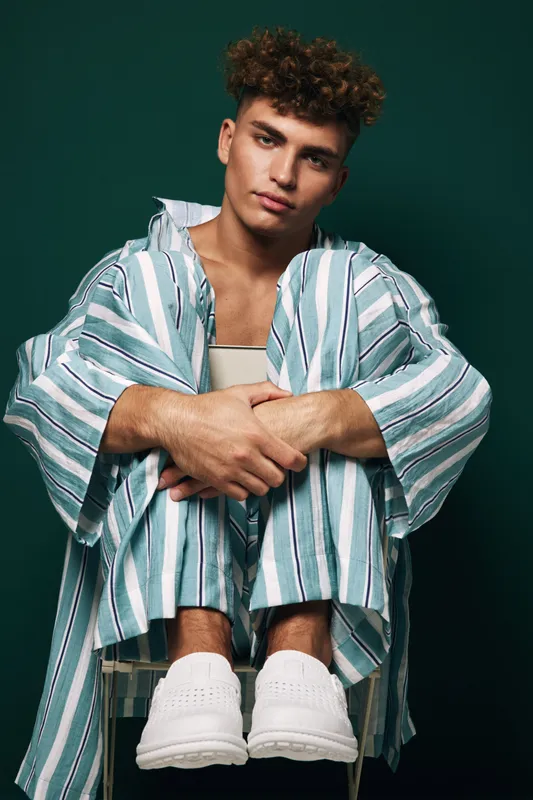 Young man with curly hair wearing striped pajamas and white shoes sitting on chair with arms wrapped around knees against dark green background in studio portrait.