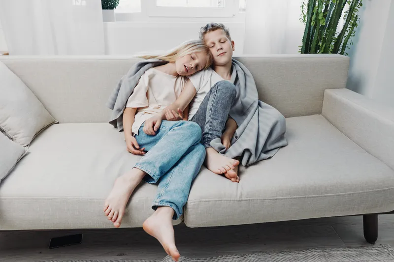 Two children sit barefoot on a beige sofa, wrapped in a gray blanket. They rest closely with eyes closed in a bright living room. Concept of comfort, warmth, sibling bond, and relaxation.