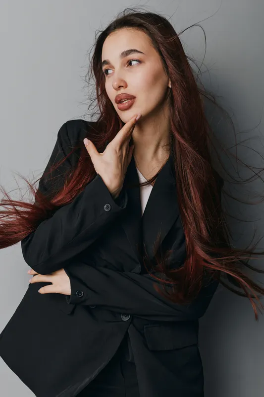 Young woman with long hair wearing black blazer posing with hand near face against gray background. Female model with natural makeup and confident expression in studio portrait.