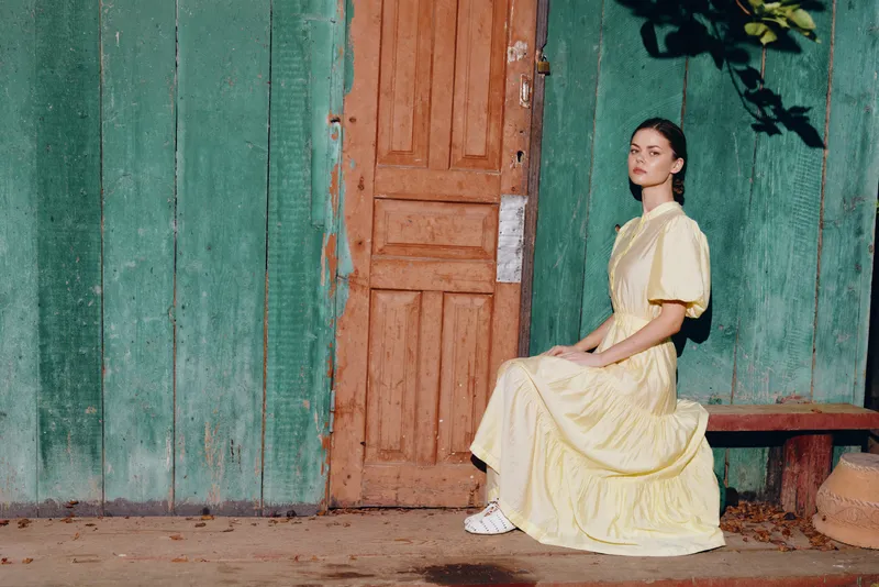 Woman seated in a yellow dress sits on a wooden bench beside a weathered green wall and doorway, capturing a calm outdoor portrait with rustic charm.