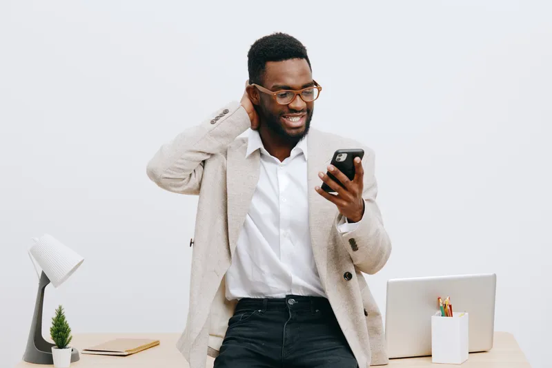 Smiling man using smartphone in bright modern office workspace with laptop and small plant, casual professional attire, happy mood, contemporary interior, natural lighting, lifestyle concept