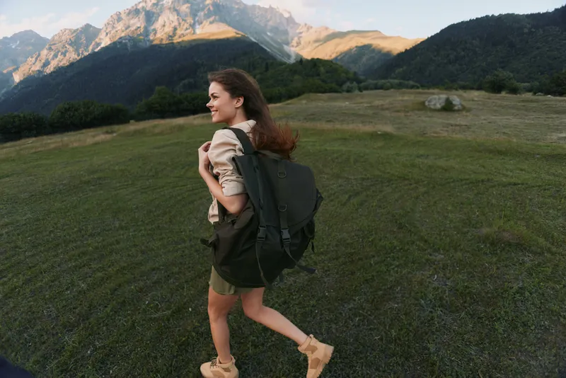 Cheerful woman outdoors, hiking with a backpack, grassy field and distant mountains in the background, sunlight on her hair, relaxed stride, natural colors and breeze