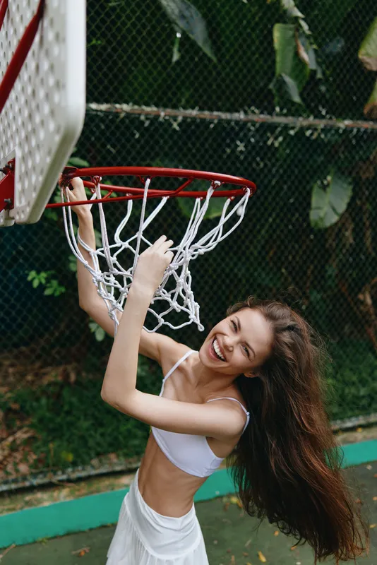 Joyful young woman playing basketball on an outdoor court, wearing a white outfit, showcasing excitement and a vibrant atmosphere amid lush green background