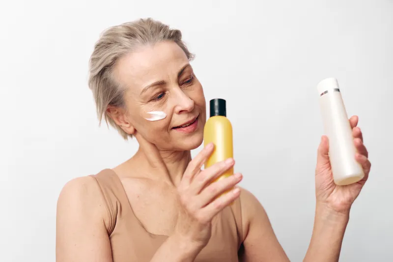 Mature woman with skincare cream on face examines two cosmetic bottles on white background. Concept of anti aging routine, self care, and beauty product choice for healthy skin.