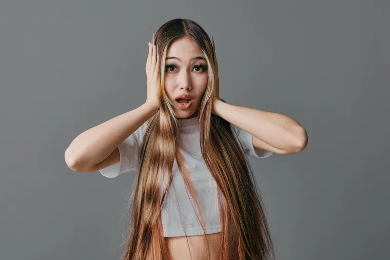 Young woman with long hair and braids holding head with surprised expression, wearing casual top against plain background for emotional portrait and fashion style.
