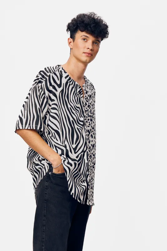 Young man wearing a stylish zebra print shirt and black pants with hands in pockets, standing confidently against a plain white background in a casual pose.