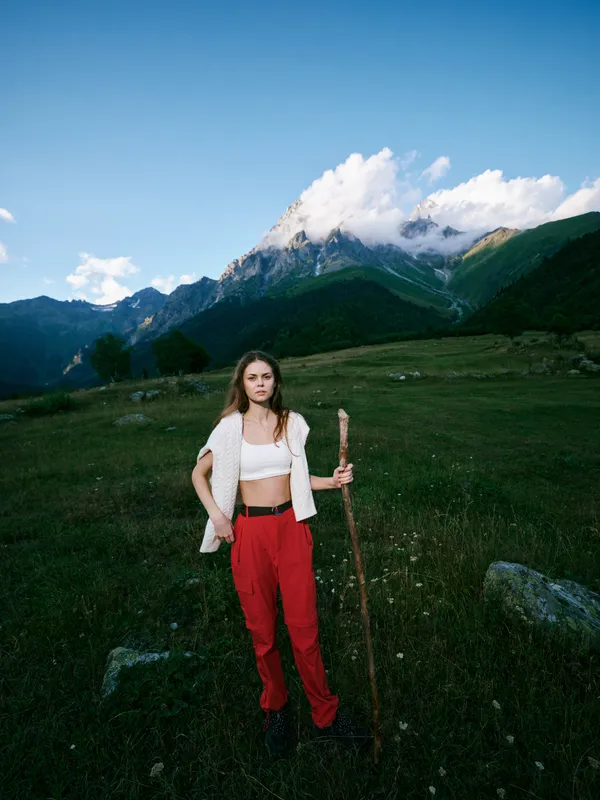 Hiking woman stands in alpine meadow with a walking stick, dramatic mountains in the background under blue sky, casual outdoor attire for adventure, nature exploration and scenic trekking in open