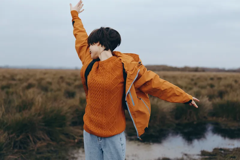 Woman lifestyle outdoor nature autumn sweater field sky river joy, young woman in orange knit waves arms in countryside beside a river, cloudy morning, calm scenic travel mood