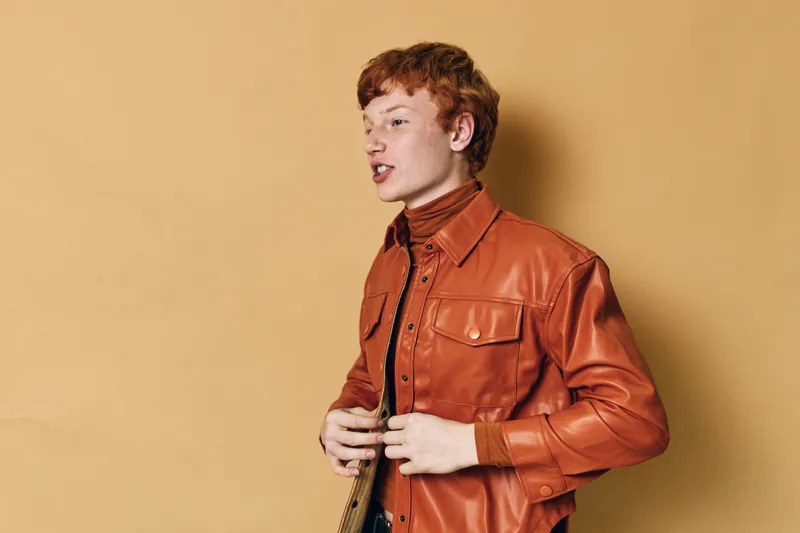 Young man with red hair wearing a brown leather jacket and turtleneck sweater standing against beige background posing in studio.