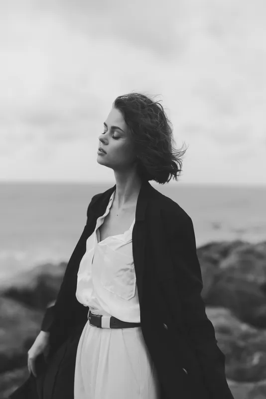 Moody coastal scene with a woman in a dress and coat, wind-swept hair, standing along the shore, monochrome tones, elegant silhouette and calm atmosphere