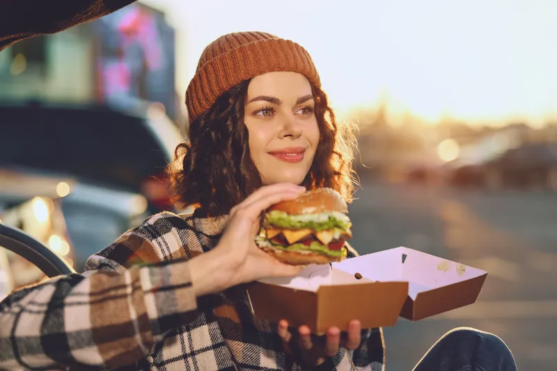 Young woman holding burger and box by car outdoors at sunset, warm smile and beanie capturing authenticity, candid lifestyle and golden hour glow for mindful living and emotional storytelling.