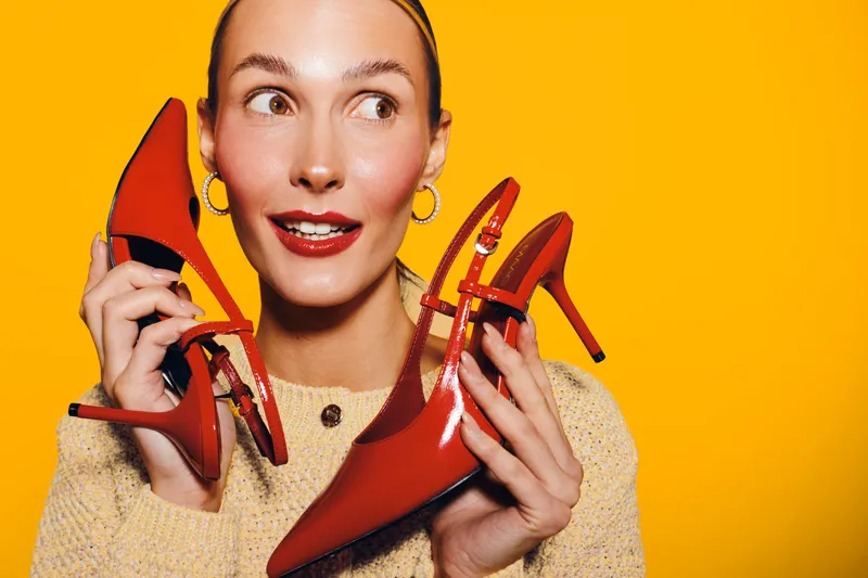 Young woman with red high heels holds shoes near face on bright yellow background. Concept of fashion, style, shopping, and trendy accessories for women.