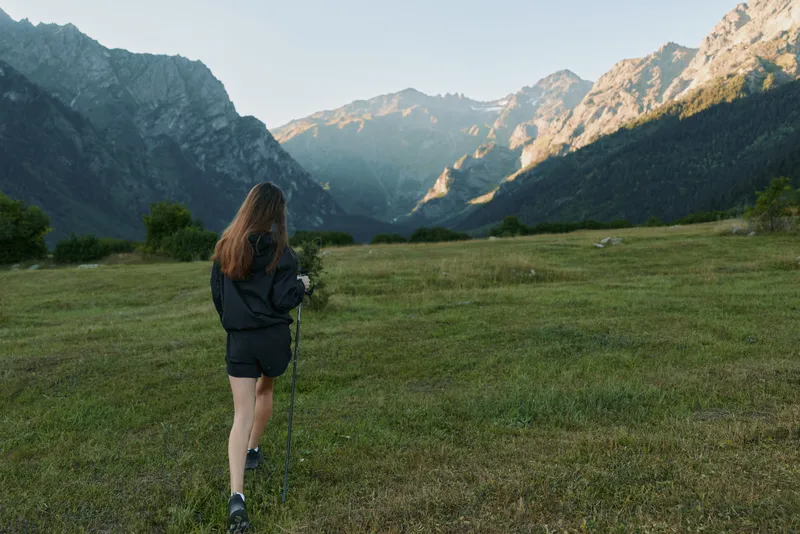 A lone hiker traverses a lush valley toward majestic mountains under a clear sky. She wears a dark jacket, backpack, and trekking poles, enjoying a serene outdoor moment.