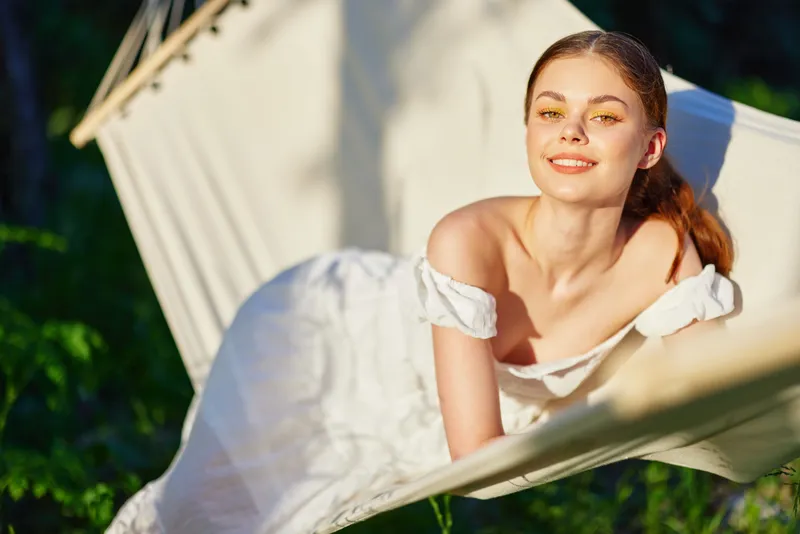 A young woman in a white dress relaxing in a hammock, enjoying the warm sunlight in a lush green garden, evoking feelings of peace and tranquility