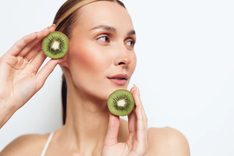 Young woman holding two kiwi halves near her face, clear skin, natural makeup, white background, fresh fruit, healthy lifestyle, bright lighting, close up portrait.