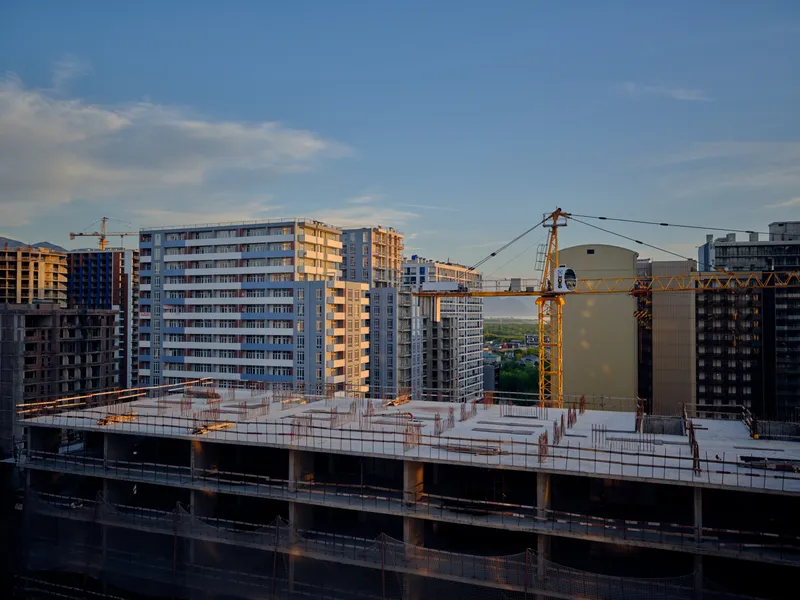 Construction site with crane next to a partially finished building in the city, apartment complex under development with concrete floors, scaffolding, workers and sunset light.