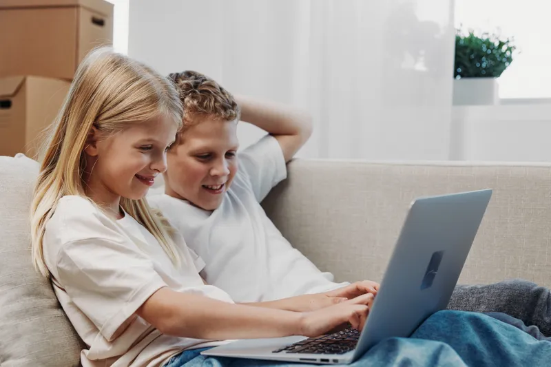 Two happy children sit on a couch using a laptop together in a bright room. Concept of learning, technology, friendship, and digital education for kids at home.