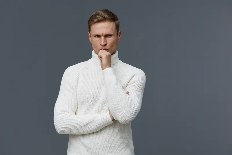 Thoughtful man in white sweater with hand on chin looking at camera serious expression indoor studio shot gray background concept thinking pondering.