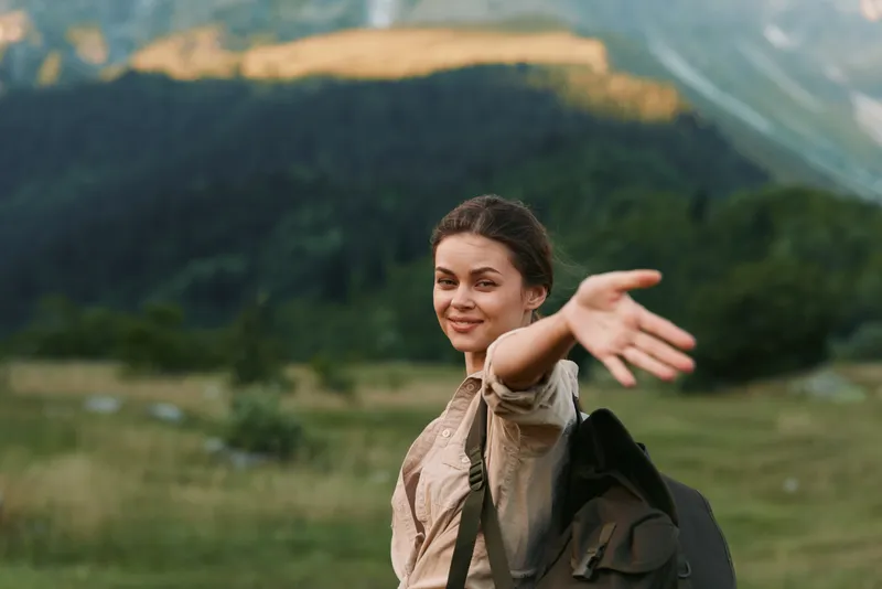 Young woman traveler with backpack smiles and reaches out hand inviting to join her in scenic mountain landscape with green meadows and forest background.