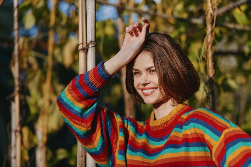 Woman smiling in a colorful rainbow sweater with a short bob haircut, leaning on a wooden post outdoors in a sunlit garden, relaxed confident mood for lifestyle portrait.
