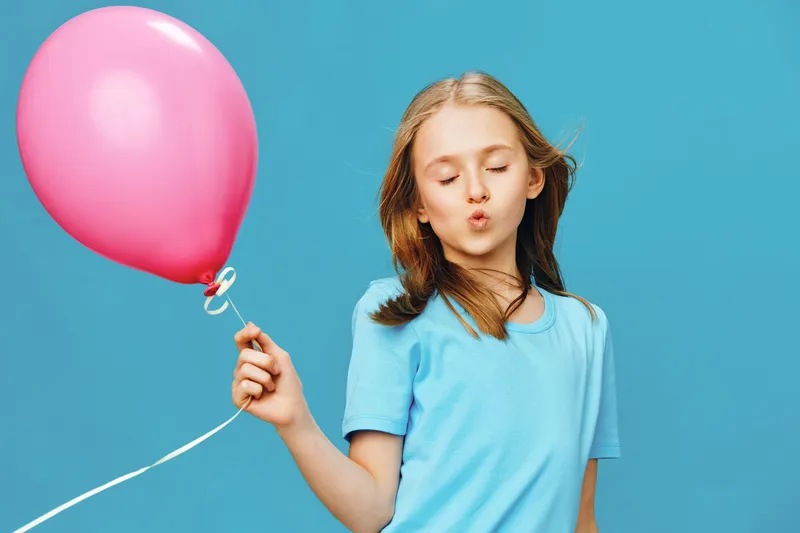 Happy girl with a pink balloon, dressed in a blue shirt, against a bright blue background, exuding joy and playfulness in a vibrant setting