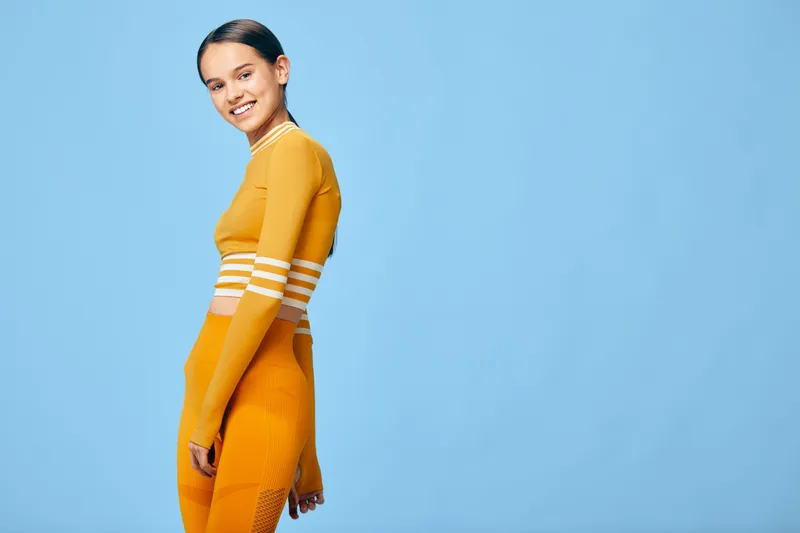 Female fitness athlete in bright orange athletic set posing in a studio with blue background, showcasing sporty wear, confident stance and active lifestyle for health and training visuals