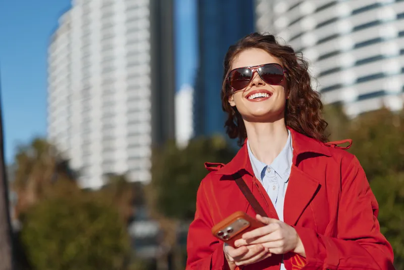 A stylish woman in a bright red coat smiles outdoors, checking her phone under sunny skies, with modern high rise buildings in the background, conveying urban fashion.