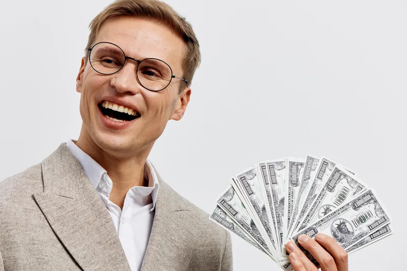 Cheerful businessman holding a fan of hundred-dollar bills, dressed in smart suit and glasses, smiling broadly against a plain white background. Concept: wealth, success, money, finance, celebration, 