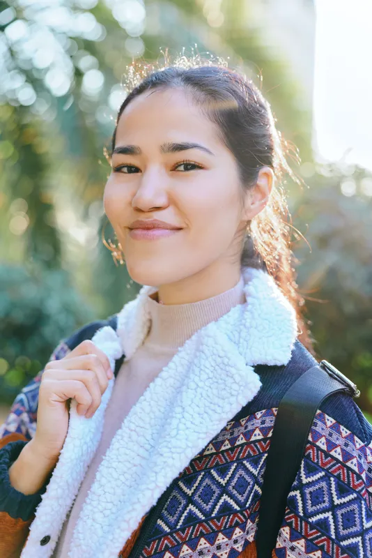 Smiling woman outdoors with backpack, patterned jacket and cozy sweater, sunlight on greenery, casual lifestyle scene conveying youth energy and travel readiness