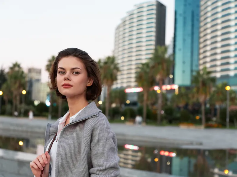 A poised woman stands outdoors near a waterfront, with modern skyscrapers and palm trees in the background, conveying a confident, contemporary urban vibe.