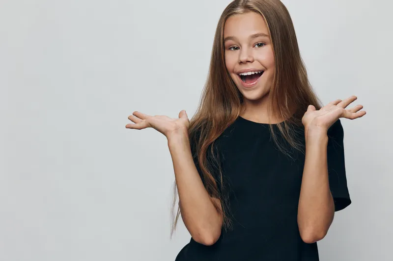 Girl with long hair wearing black shirt, smiling and posing with open hands on light background. Happy child with expressive face in casual clothes for lifestyle or childhood concept.
