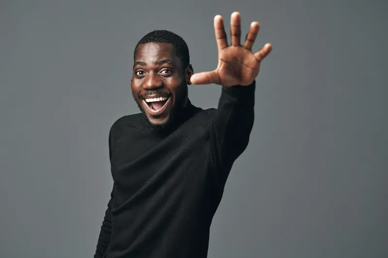 African man with dark skin smiling and reaching hand forward in creative conceptual visual style on gray background. Happy male portrait with positive expression and casual black sweater.