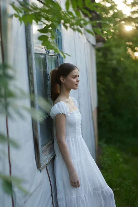 Young woman in a white dress standing by a weathered wall, evoking calmness, nature, and summer vibes, surrounded by greenery