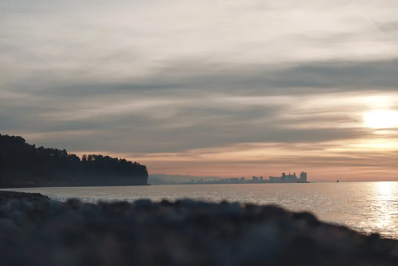 Sunset over calm water with a distant city skyline, gentle horizon and rocky shore in foreground, creating a peaceful landscape at dusk and inviting mood for travel, nature, and evening reflection.
