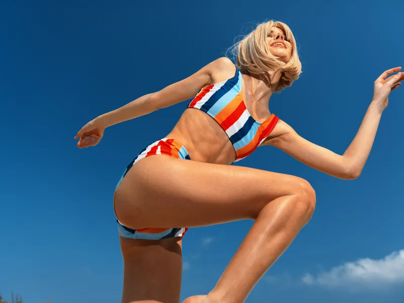Professional athletic woman mid jump in a colorful striped bikini against a clear blue sky, capturing motion, balance, and sunlit vitality in a dynamic beach scene.