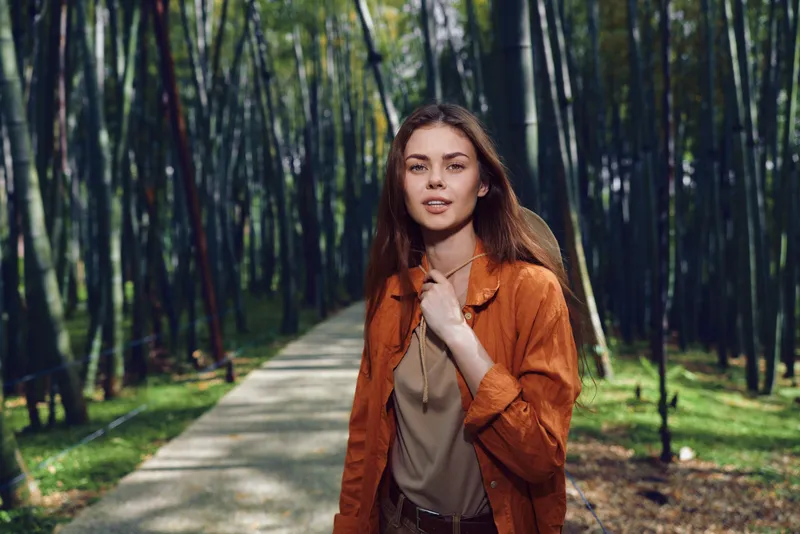 Woman portrait walking on a path through a bamboo forest, casual urban outfit, confident young female in nature, sunlight filtering through tall bamboo trees during daytime.