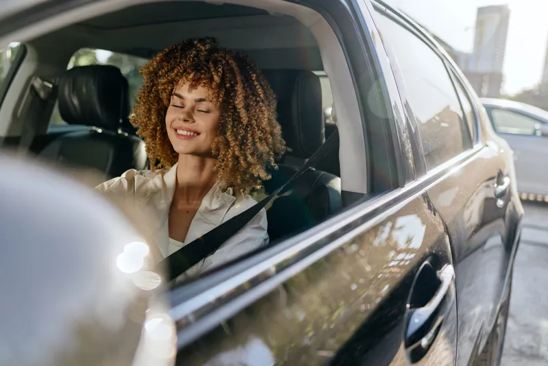 smiling woman driving car with curly hair, sunlight, casual shirt, urban background, happy, relaxed expression