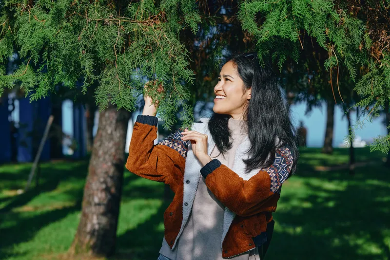 Outdoor scene featuring a smiling woman in a cozy cardigan reaching toward vibrant leaves, enjoying shade, sunlight, and fresh air in a tranquil park with trees, grass, and light breeze