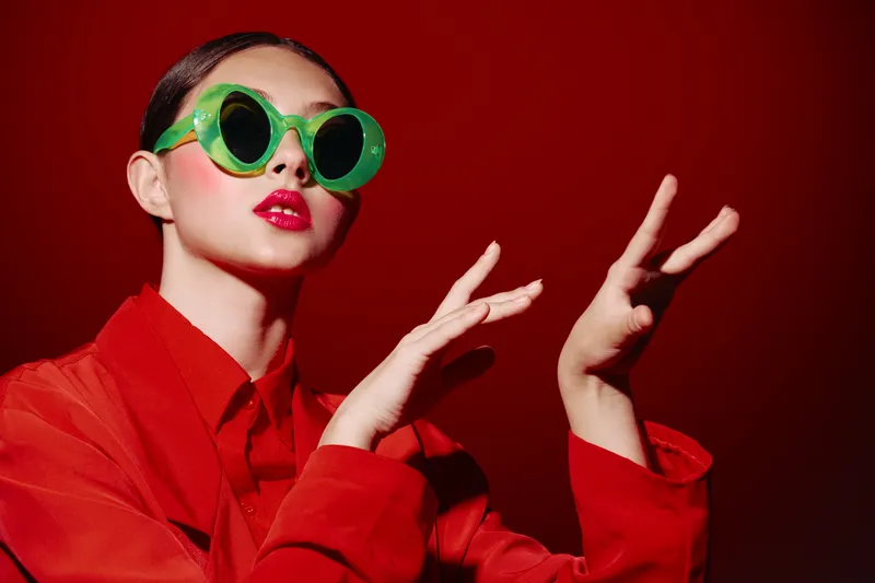 Fashion model with bold green eyewear and bright red lipstick poses with expressive hands wearing a matching red outfit against a red background. Creative beauty concept.